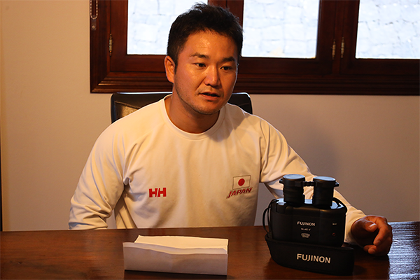 A short-haired man is sitting in a chair, answering questions in an interview. Binoculars are placed on the desk in front of him.